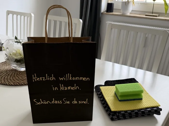 Schwarze Papiertüte auf dem Esstisch, von Hand beschriftet mit „Herzlich willkommen in Hameln. Schön, dass Sie da sind.“, daneben Spültuch und Schwamm